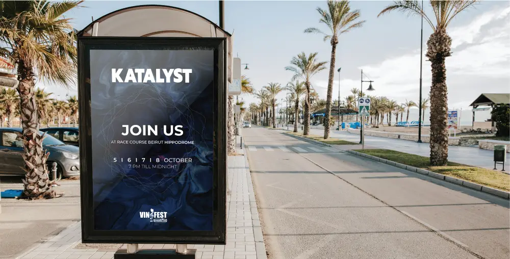 Bus stop advertisement on a coastal street with palm trees, promoting 'KATALYST.' The ad features the text: 'KATALYST. JOIN US. ONE OF A KIND SUMMER EXPERIENCE. SIGN UP NOW. WWW.KATALYST.COM.' The background shows a clear sky and ocean view.