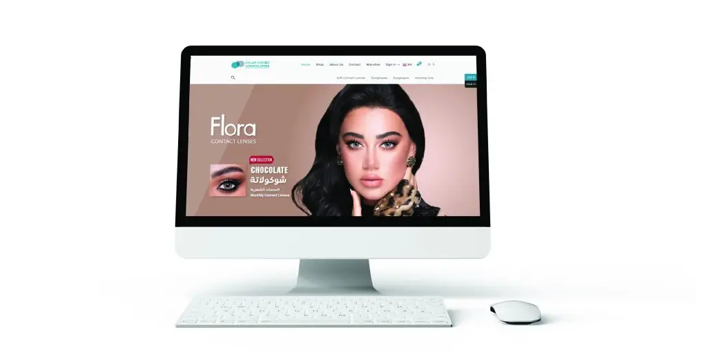 Computer monitor displaying a website related to beauty or cosmetics, featuring a large image of a person's eyes and hair, with the text 'Flora Lash & Brow.' A promotional badge says 'NEW ARRIVAL' and '50% OFF.' The website has a soft pink background and a clean, modern design. The monitor is accompanied by a keyboard and a mouse, all set against a white background.