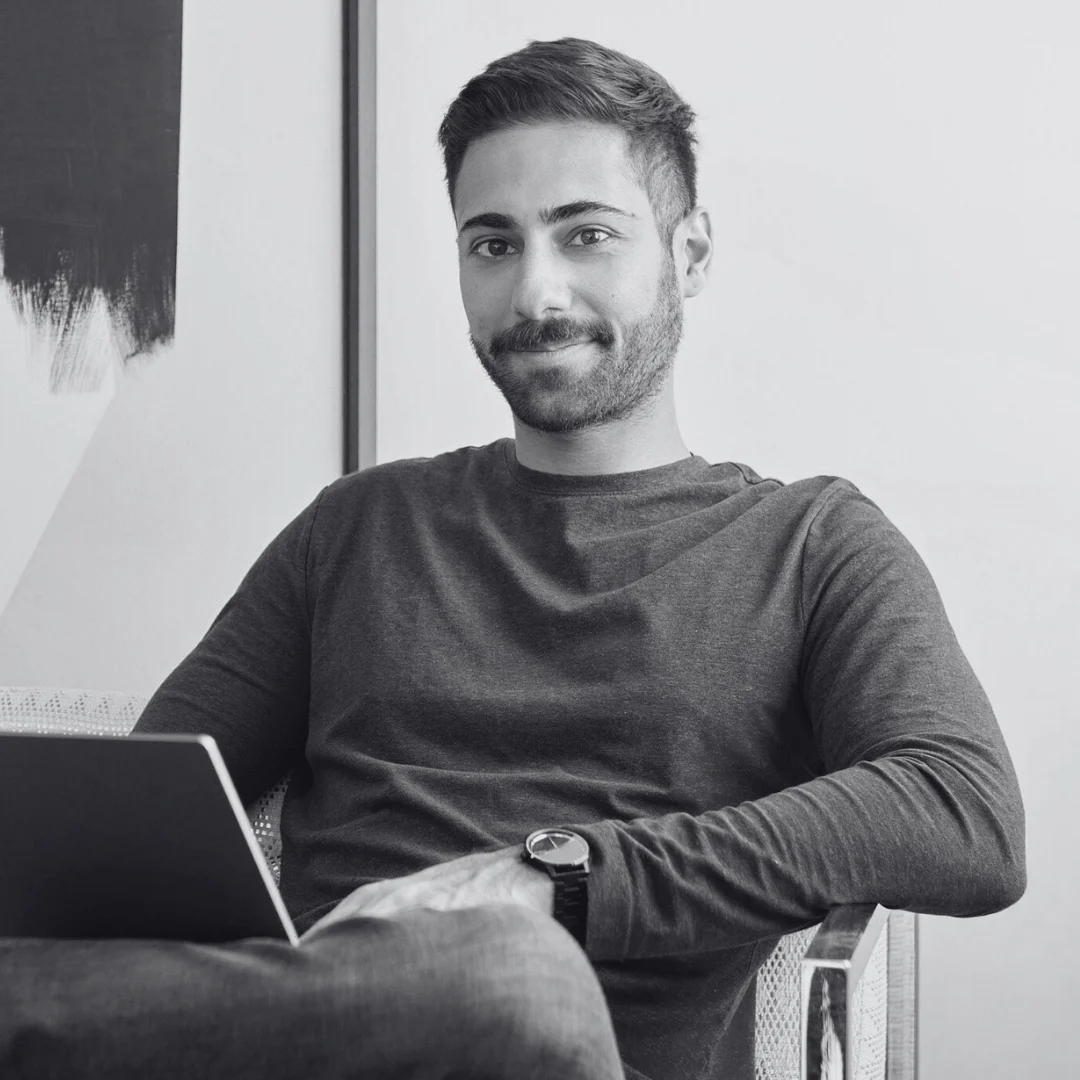 Person sitting on a chair with legs crossed, holding a laptop. The individual is wearing a long-sleeved shirt and a wristwatch. The background includes a wall with abstract art. The image is in black and white.