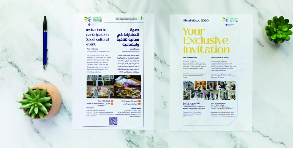 Two printed flyers placed on a marble surface, with a pen and two small potted plants nearby. The flyer on the left is in Arabic and English, with a headline, sections of text, a QR code, and images of people and activities. The flyer on the right, in English, reads 'Your Exclusive Invitation' and includes sections of text and images of people and activities.