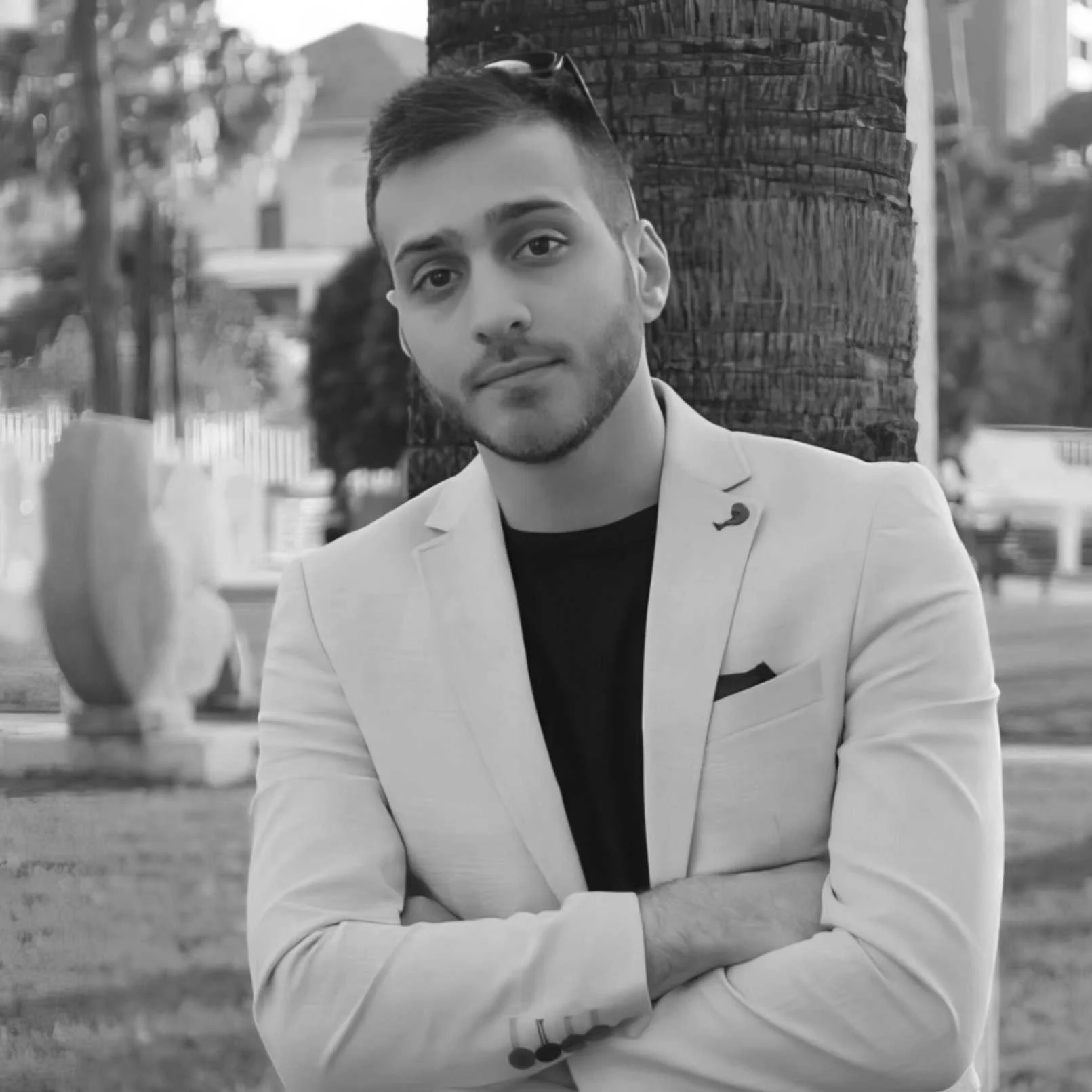 Person wearing a light-colored blazer over a dark shirt, standing in front of a tree trunk in an outdoor setting. The blazer has a pocket square and a small pin on the lapel. The background includes trees and some architectural elements.